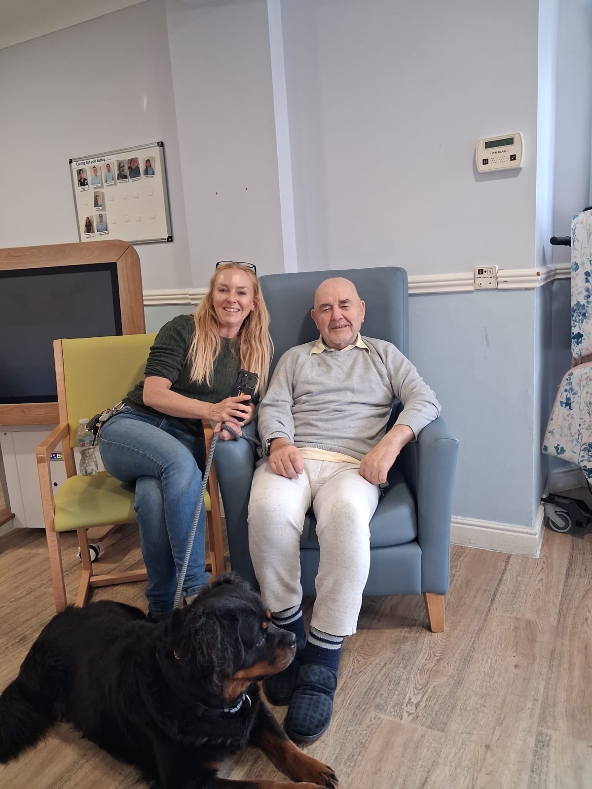 Jo sitting beside her dad in his care home lounge, with Ida the Rottweiler lying at their feet
